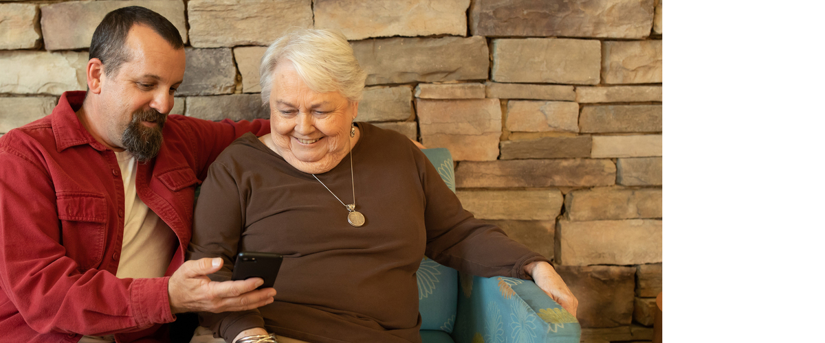 A man and an older woman sitting on a couch with wide smiles looking at a smartphone that the man is holding