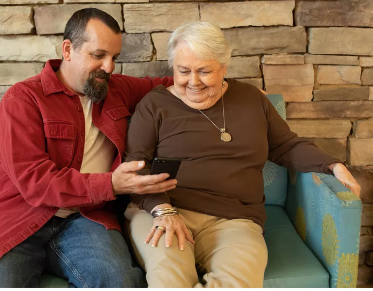 A man and an older woman sitting on a couch with wide smiles looking at a smartphone that the man is holding