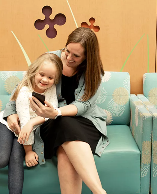 A woman holding her young daughter while they both smile and look at the smartphone she's holding